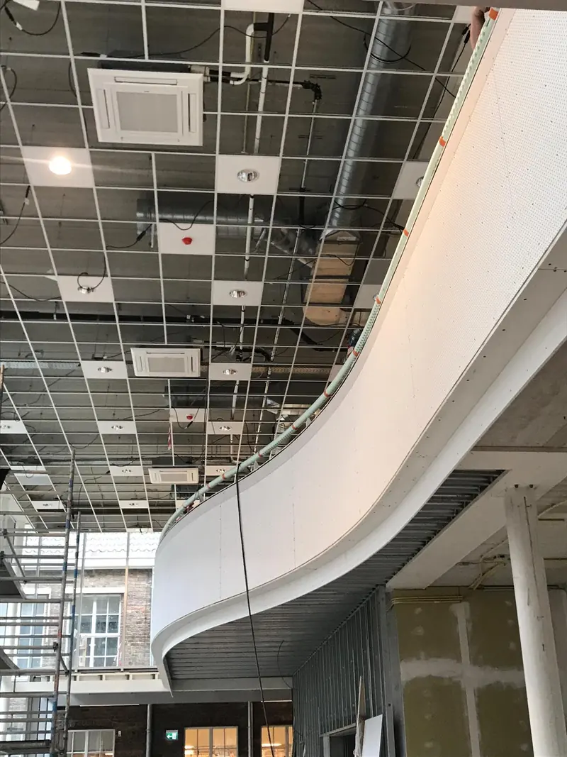 Gebogen vide-rand in metal stud onder een open plafondraster, tijdens de bouw